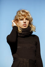 Load image into Gallery viewer, A blonde model pushing her hair behind her ear, wearing a black outfit  with a textured scarf accessory, against a blue sky 