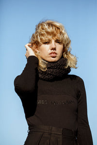 A blonde model pushing her hair behind her ear, wearing a black outfit  with a textured scarf accessory, against a blue sky 