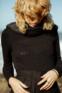 blonde model looking down, wearing a branded meg taljaard, black jersey with high waisted trousers and a black textured scarf accessory