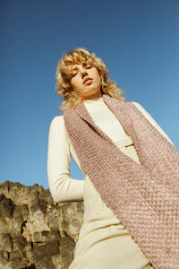 Blonde female model against a blue sky, looking down, wearing a dirty pink, textured scarf accessory