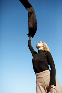 fashion model wearing a black jersey and beige pants throwing a scarf up in the air 