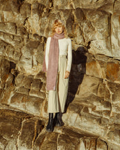 Load image into Gallery viewer, A blonde female model standing against a rocks, wearing a cream polo neck with beige trousers and a pink scarf accessory, 