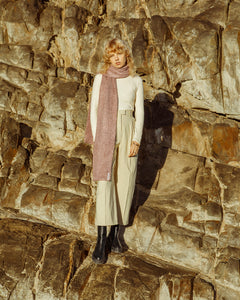 A blonde female model standing against a rocks, wearing a cream polo neck with beige trousers and a pink scarf accessory, 